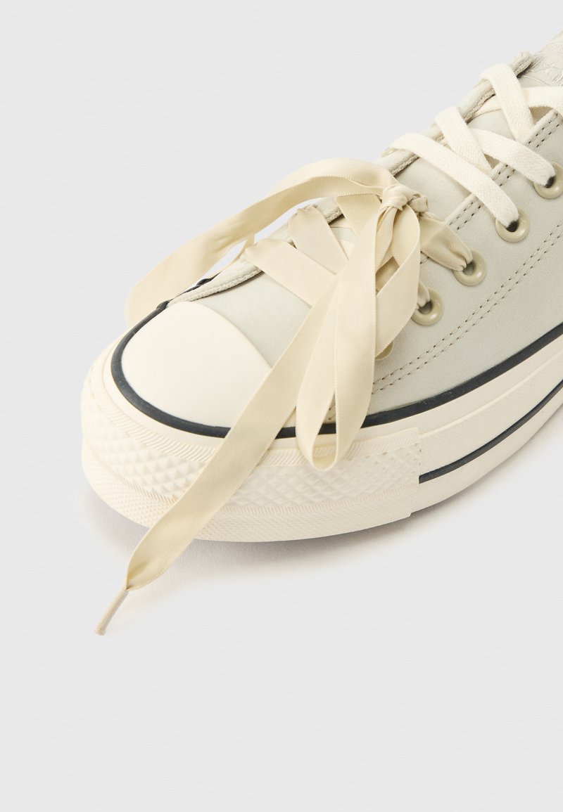 Sneaker in tela color bianco sporco con suola spessa, striscia nera e larghe stringhe di raso crema annodate a fiocco su uno sfondo grigio chiaro.