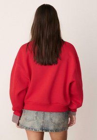 Felpa rossa oversize con spalle scese, polsini a costine e vestibilità comoda. Abbinata a una minigonna in denim con tasche posteriori.