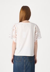 Biała koszulka z misternymi koronkowymi rękawami, widok od tyłu. Gładka tekstura i wygodne dopasowanie, w połączeniu z jeansową spódnicą.