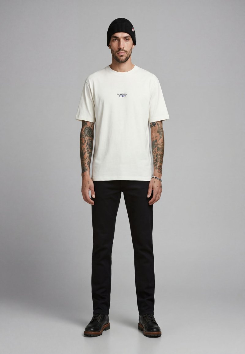 Mann mit Tattoos, der eine schwarze Beanie, ein weißes T-Shirt mit Text, schwarze Hose und schwarze Schuhe trägt, steht vor einem schlichten grauen Hintergrund.