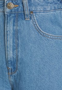 Vaaleansiniset farkut, joissa on teksturoitu pinta, messinkinen nappi vyötäröllä ja oranssit ompelutaskujen sekä saumoiden kohdalla.