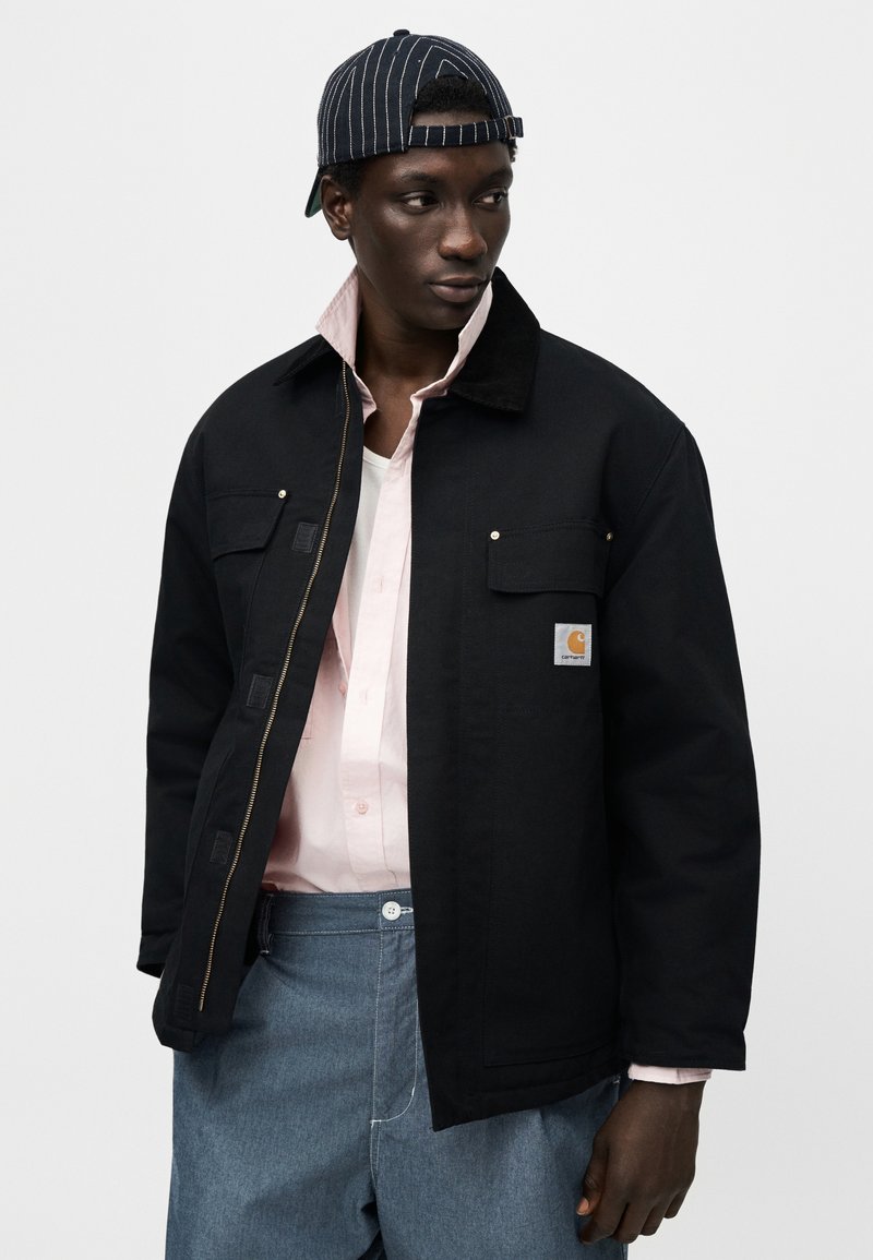 Zwarte stoffen jas met een ritssluiting aan de voorkant, twee borstzakken en een logopatch. Draagt eroverheen een roze blouse met knopen, een wit T-shirt en grijze broek.