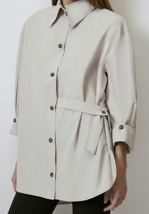 Beżowa koszula na długi rękaw, zapinana na guziki, z kołnierzykiem i wiązaniem z boku, noszona z czarnymi spodniami, pokazana od szyi do połowy uda.