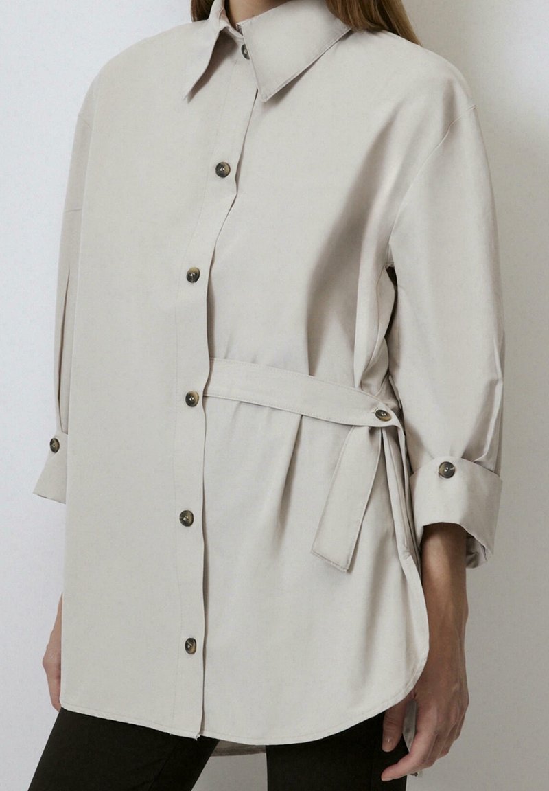 Chemise beige à manches longues avec col et détail de nouage sur le côté, portée avec un pantalon noir, visible du cou au milieu de la cuisse.