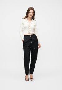 Γυναίκα στέκεται προς τα εμπρός φορώντας λευκό κομμένο ζακέτα με κουμπιά, μαύρο παντελόνι ψηλόμεσο με φερμουάρ και μαύρες γόβες.