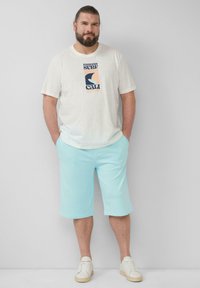 T-shirt gris clair pour homme avec un texte graphique en marine et pêche, associé à un short vert menthe et des baskets blanches, se tenant les mains dans les poches.