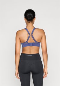 Sustentador deportivo morado con un diseño de malla en varias capas, tirantes ajustables y cierre de gancho. Combinado con leggings negros de tiro alto con un logo.