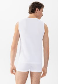 Man in een witte mouwloze shirt en witte boxer shorts, gezien van achteren tegen een effen witte achtergrond.