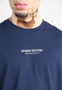 Navy zila T-kreklu, kas izgatavots no kokvilnas, ar apaļu kakla izgriezumu. Priekšdaļā ir balts teksts ar uzrakstu "STUDIO EDITION ALPHA INDUSTRIES INC."