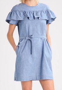 Robe en denim bleu clair avec un col volanté, des manches courtes, une taille à nouer et des poches sur les côtés. Design simple et ajusté avec une texture lisse.