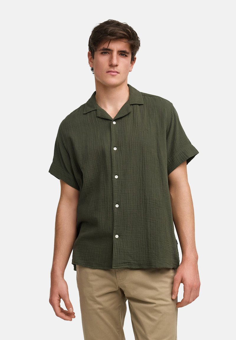 Chemise à manches courtes, couleur olive, en tissu texturé, avec un col et des boutons sur le devant, associée à un pantalon beige clair.