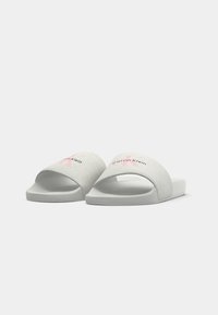Un paio di pantofole grigie chiare Calvin Klein con logo "CK" rosa e testo nero su fasce larghe, su uno sfondo bianco.