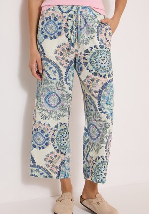 Frau trägt eine Hose mit weitem Bein mit mehrfarbigen Kreis- und Blumenmustern, ein hellrosa Oberteil und beige Schlupfschuhe.