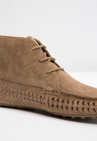 Bottine en daim marron clair avec fermeture à lacets sur le devant et une couture en zigzag détaillée le long de la semelle et du côté supérieur.