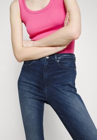 Top de tirantes de color rosa con textura acanalada, combinado con vaqueros de mezclilla oscuros de tiro alto; textura suave, diseño ajustado, con bolsillos frontales y cierre de botón.