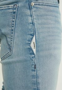 Jeans de mezclilla azul claro con un ajuste slim, costuras visibles y una etiqueta de marca en el costado. La tela parece suave con una ligera textura.