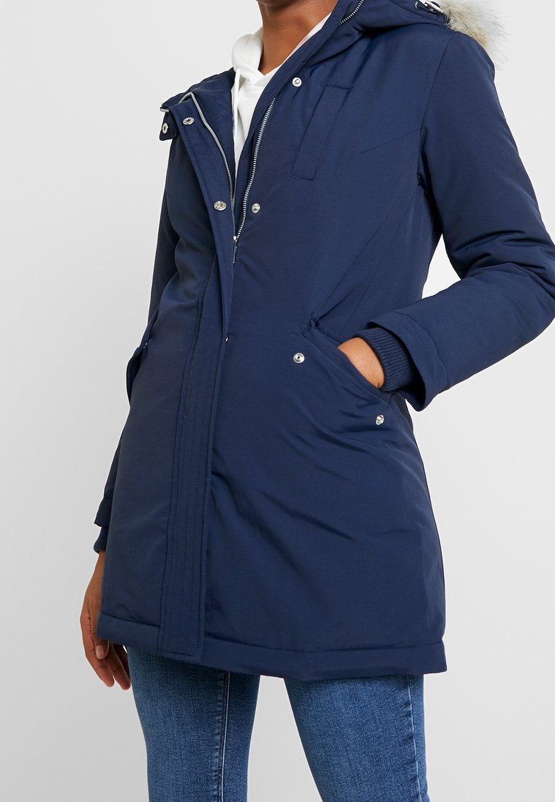 Navy blue parka jacket with a fur-trimmed hood, zip and snap button closure, front pockets, and ribbed cuffs. Features a smooth, textured fabric.