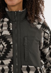 Veste en polaire avec un motif géométrique noir et gris, dotée d'un col montant, d'une fermeture éclair et d'une poche poitrine pour le rangement.