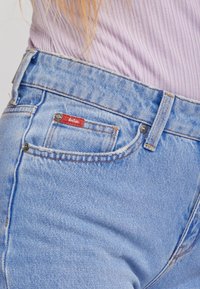 Jeans de mezclilla azul claro con una cinturilla acanalada, que presentan una etiqueta roja, cierre de botón metálico y un sutil desgaste en la tela.
