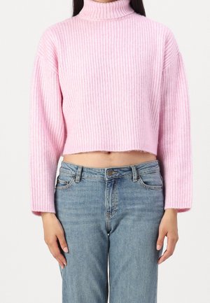 Vrouw draagt een lichtroze geribbelde cropped coltrui en hoogtalgige blauwe spijkerbroek met handen ontspannen langs haar zij.