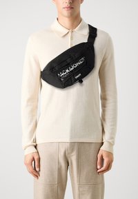 Black nylon sling bag featuring a prominent white logo, zippered pocket, and adjustable strap, worn across the chest over a beige sweater.