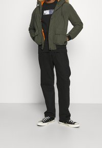 Giacca a cappuccio verde oliva con rifiniture in pelliccia, fodera arancione e due tasche frontali, abbinata a una maglietta nera, pantaloni neri e sneakers nere.