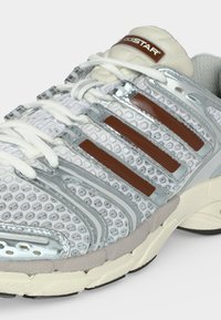 Λευκό και ασημί αθλητικό παπούτσι με καφέ ρίγες, υφασμάτινο επάνω μέρος με υφή, λευκά κορδόνια και μαξιλαρωμένη σόλα για αθλητική χρήση.