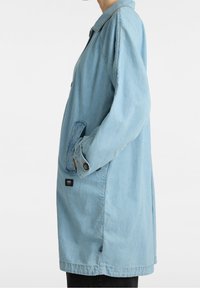 Vaaleansininen farkkutakki, jossa on kaulus, pitkät hihat ja sivutaskut. Hihansuissa on napit ja helman lähellä merkintälappu.