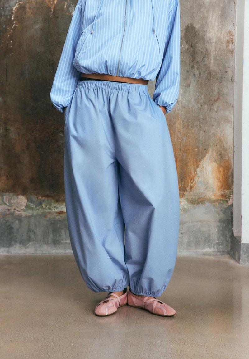 Personne portant un pantalon bleu clair ample et une veste courte assortie, debout sur un sol poli devant un mur texturé.