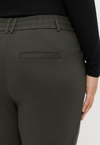 Tmavě zelené kalhoty s elastickým pasem a detailní plochou zadní kapsou. Hladká textura, přiléhavý střih a vysoký pas.