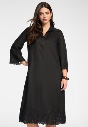 Frau trägt ein schwarzes knielanges Kleid mit dreiviertel Ärmeln und Spitzenbesatz an den Bündchen und am Saum, posiert mit einer Hand in der Hüfte.