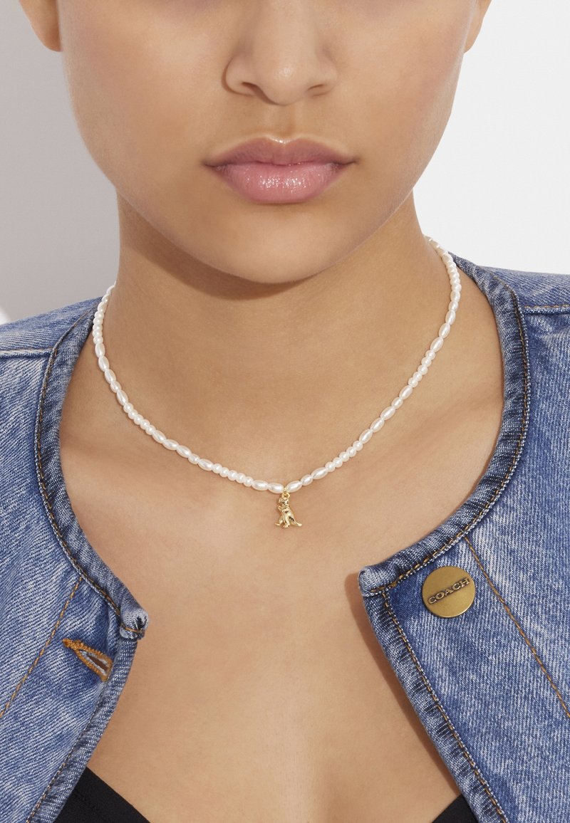 Halskette aus weißen Perlen mit einem zentralen goldenen Anhänger. Getragen über einer Jeansjacke, die die glatte Textur und die runden Formen der Perlen zur Schau stellt.