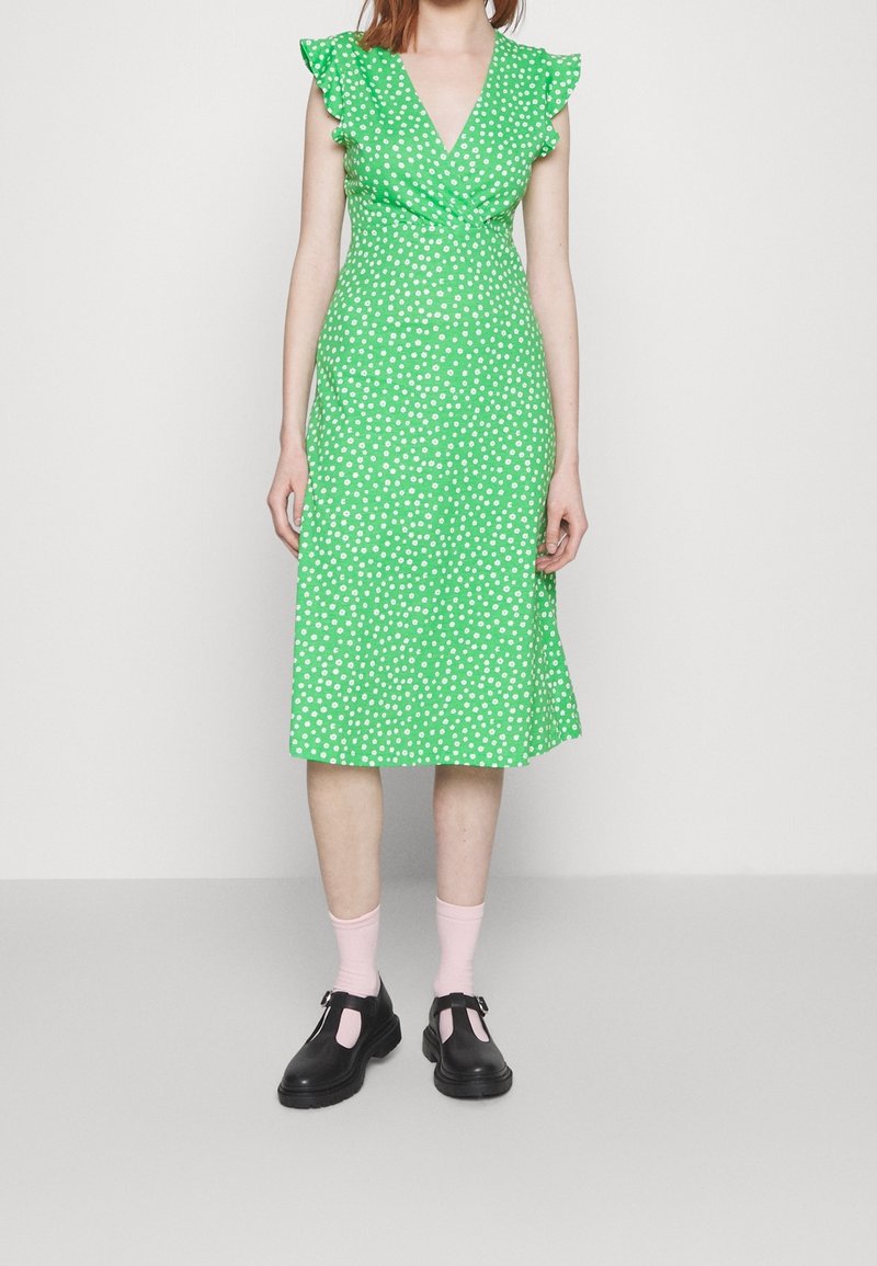 Robe midi verte à pois avec des manches volantes, design enroulé et ourlet évasé. Associée à des chaussures noires et des chaussettes roses.