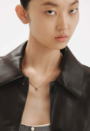 Chaqueta de cuero negra con diseño de cuello. Un delicado collar de oro presenta un colgante rectangular con acentos rojos y verdes.