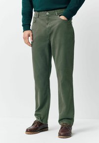 Mees, kes kannab oliivirohelisi sirgeid pükse eesmine kottide ja pruunide nahksete nööbisaabastega, seisab, hoides ühte kätt taskus.