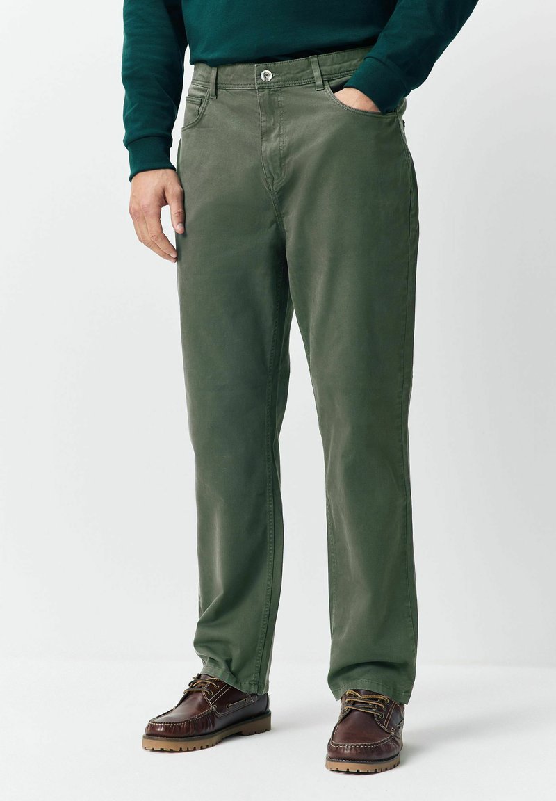 Mees, kes kannab oliivirohelisi sirgeid pükse eesmine kottide ja pruunide nahksete nööbisaabastega, seisab, hoides ühte kätt taskus.