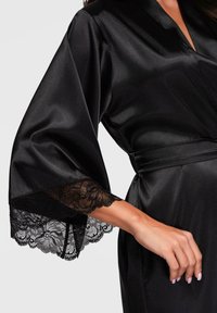 Donna che indossa un accappatoio di raso nero con ampie maniche caratterizzato da un bordo in pizzo floreale, legato in vita, che mostra il suo braccio e mano destri.