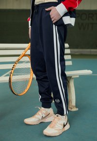 Jūrų laivyno arbatos spalvos sportinės kelnės su baltomis šoninėmis juostomis, elastinga juosta ir kišenėmis. Dėvimos su baltais ir rožiniais Nike sportbačiais, laikant oranžinį teniso raketę.
