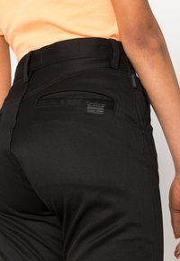 Detailní záběr na osobu, která má na sobě černé kalhoty G-Star RAW a oranžové tričko, přičemž je vidět zadní kapsa a část jejího ramene.