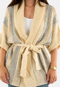 Cardigan a maglia aperta in beige e grigio con cintura. Presenta una trama spessa e una leggera brillantezza lungo le strisce. Maniche corte, vestibilità comoda.