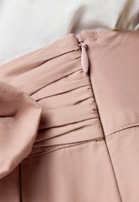 Prenda de tela rosa con detalles plisados y cierre de cremallera. Textura suave y acabado liso. Presenta un adorno de lazo cerca de la cintura.