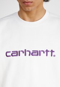 Sweatshirt blanc avec un grand logo "carhartt" brodé en violet sur la poitrine, doté d'un col rond côtelé.