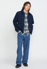 Chaqueta azul marina con cierre de botones y dos bolsillos frontales, llevada sobre una camisa de cuadros y combinada con pantalones de mezclilla azules y zapatos negros.