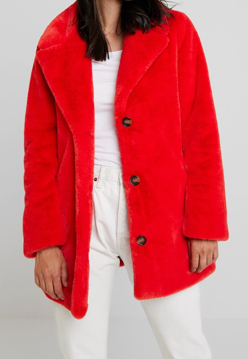 Abrigo de pelo sintético en vibrante rojo con cuello solapa, cuatro botones grandes y mangas largas, combinado con pantalones blancos. Textura suave.