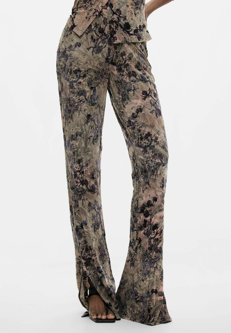 Mujer que lleva pantalones de talle alto y pierna ancha con patrón floral abstracto beige y negro y aberturas frontales, combinados con tacones negros abiertos en la punta.