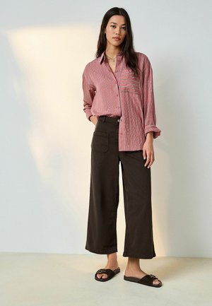 Woman wearing a loose red striped button-up shirt with a chest pocket, black wide-leg cropped pants, and black slide sandals.