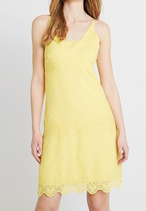 Kvinde iført en gul, ærmeløs kjole med blonder langs kanten og over knæet, stående mod en ensfarvet baggrund.