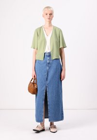 Lys grønn, kortermet bluse kombinert med et langt denimskjørt med en splitt foran. Modellen holder en liten brun håndveske. Nøytrale sko.