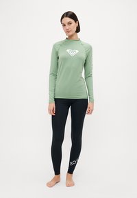 Langermet lysegrønn rash guard med hvitt logo og detaljer, kombinert med svarte leggings med hvit "ROXY" tekst. Glatt tekstur.