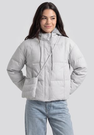 Frau trägt eine hellgraue gesteppte Jacke und hellblaue Jeans mit einer passenden kleinen Umhängetasche, lächelnd und zur Seite schauend.